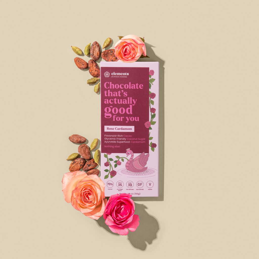A chocolate bar package from elements wellness, labeled 'Rose Cardamom', styled on a beige background with pink roses and cocoa beans arranged around it.