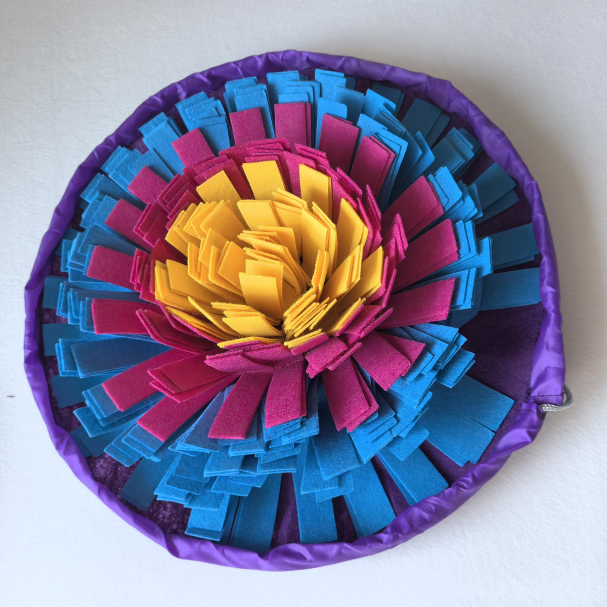 A circular fabric pouch with a colorful layered flower-like design, featuring yellow, magenta, and blue petal-like layers around a purple border, photographed on a plain light background.
