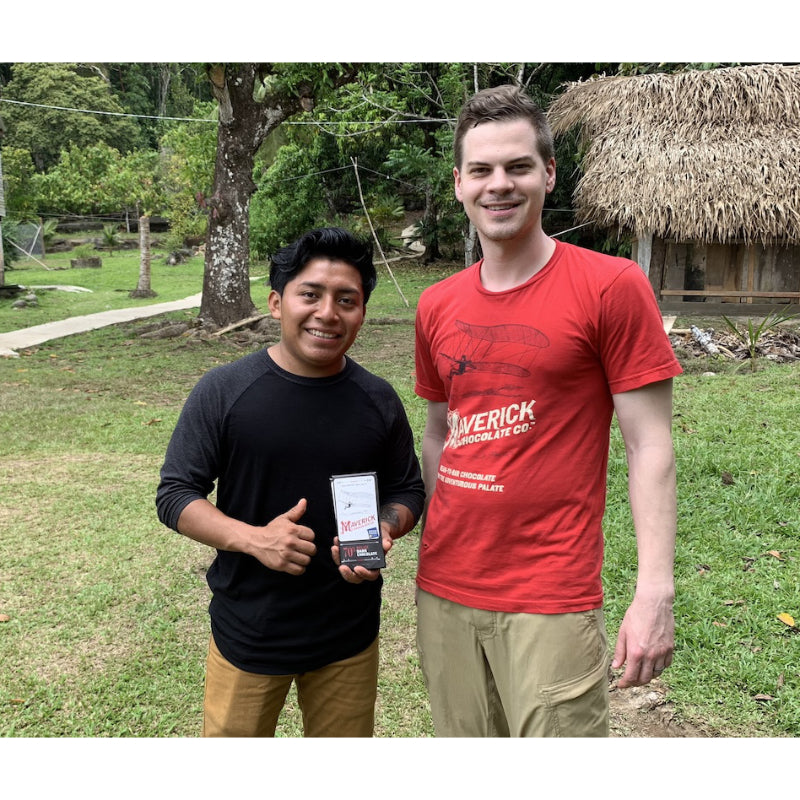 Maverick Chocolate Co. team member with a cacao farmer in Belize holding the 70% dark chocolate bar, outdoors by a thatched hut