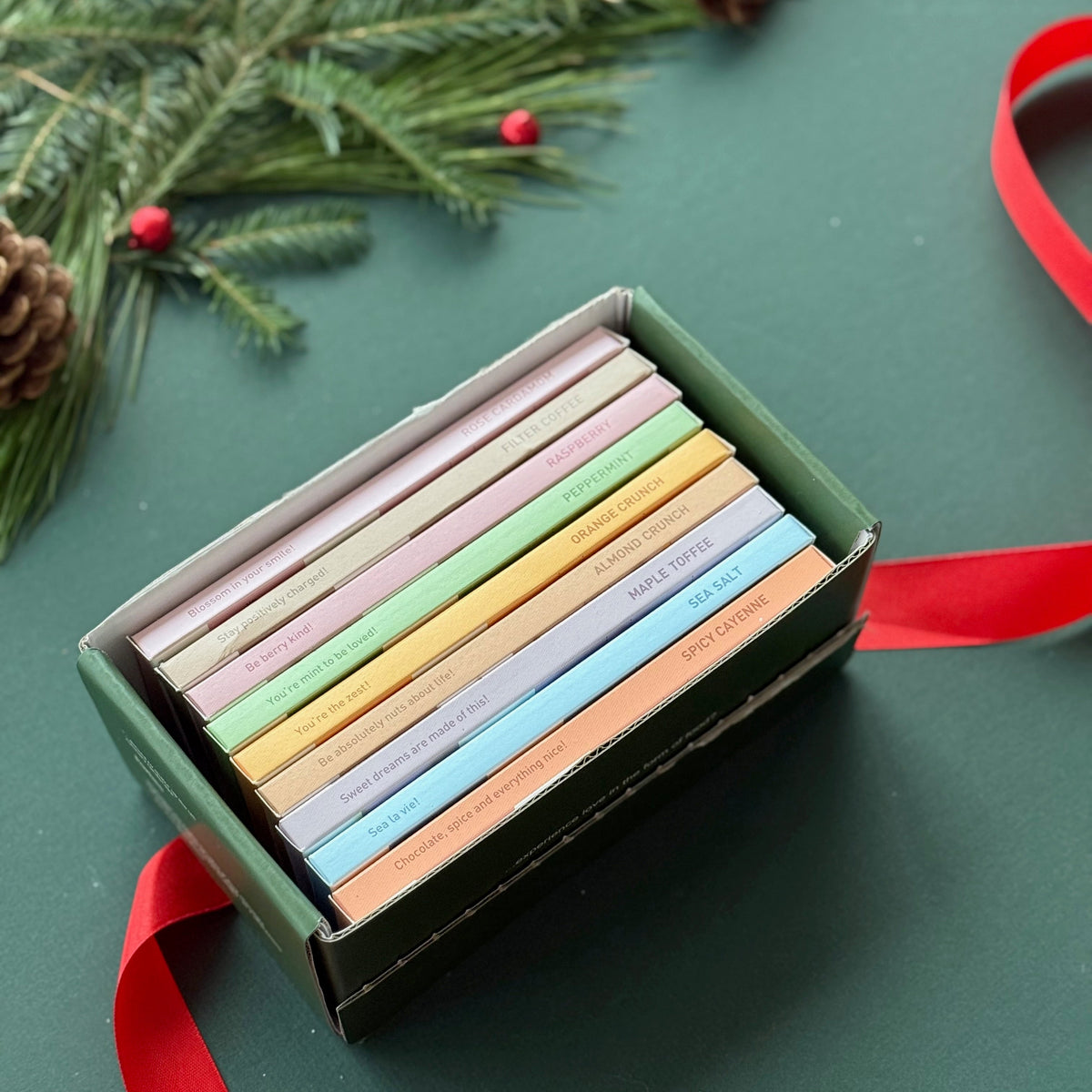 Soma collection gift box with nine pastel-wrapped 75% dark chocolate bars on a green background with holiday greenery and red ribbon